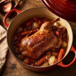 Un caneton rôti doré dans une cocotte rouge avec des carottes et des oignons sur une table rustique